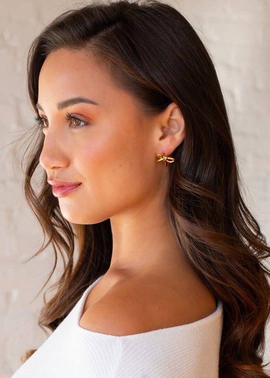 A woman with long wavy brown hair wears the Luxe Gold Bow Stud Earrings by panacheaccessories and a white off-the-shoulder top, shown in profile against a light, blurred background.