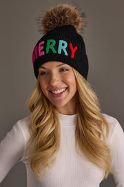 A smiling woman wears a Black Merry Knit Pom Hat, a black knit hat featuring the word MERRY in colorful, embroidered block letters and a large faux fur pom-pom on top.