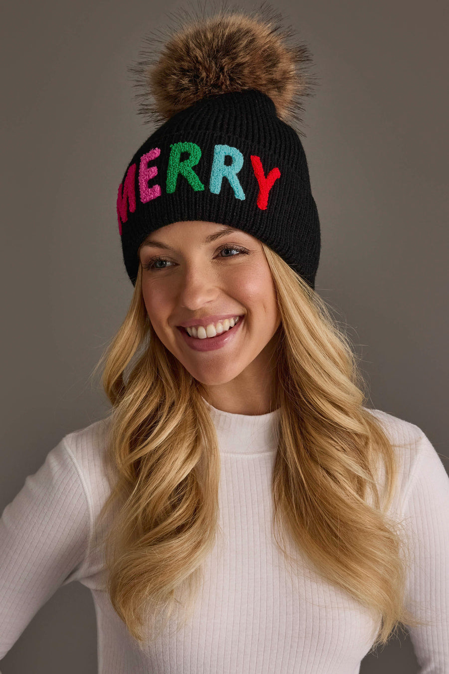 A smiling woman wears a Black Merry Knit Pom Hat, a black knit hat featuring the word MERRY in colorful, embroidered block letters and a large faux fur pom-pom on top.
