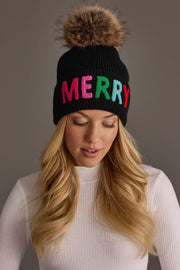 A Panache Apparel Collection Black Merry Knit Pom Hat featuring a fleece-lined black knit, a natural faux fur pom-pom, and the word MERRY stitched in colorful, multi-toned letters.