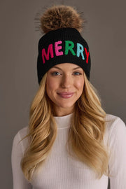 A woman wears a Black Merry Knit Pom Hat, a fleece-lined, black knit pom hat featuring a natural faux-fur pompom and the word MERRY stitched in colorful letters across the front.