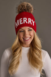 A smiling woman wears a red Red Merry Knit Pom Hat by Panache Apparel Collection, featuring the word MERRY in white letters and a large, fluffy pom-pom on top.