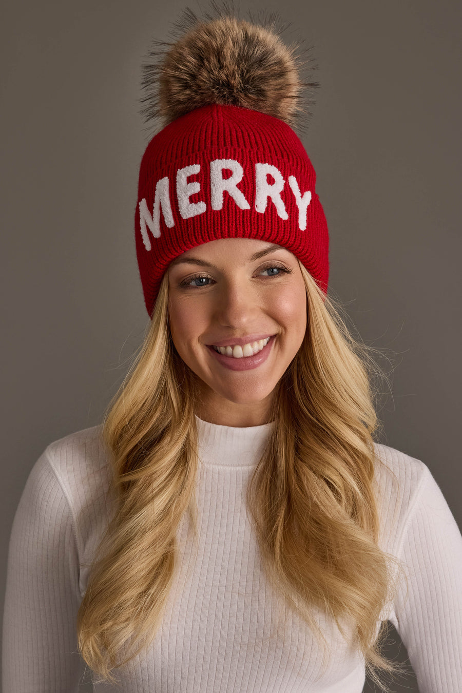 A smiling woman wears a red Red Merry Knit Pom Hat by Panache Apparel Collection, featuring the word MERRY in white letters and a large, fluffy pom-pom on top.