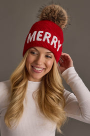 A smiling woman wears a Red Merry Knit Pom Hat in red knit with the word MERRY in white letters and a large, fluffy pom-pom on top.