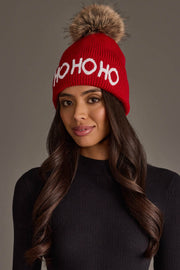 A person wears a Red Ho Ho Ho Knit Pom Hat with Ho Ho Ho in white letters and a large faux fur pom-pom on top.