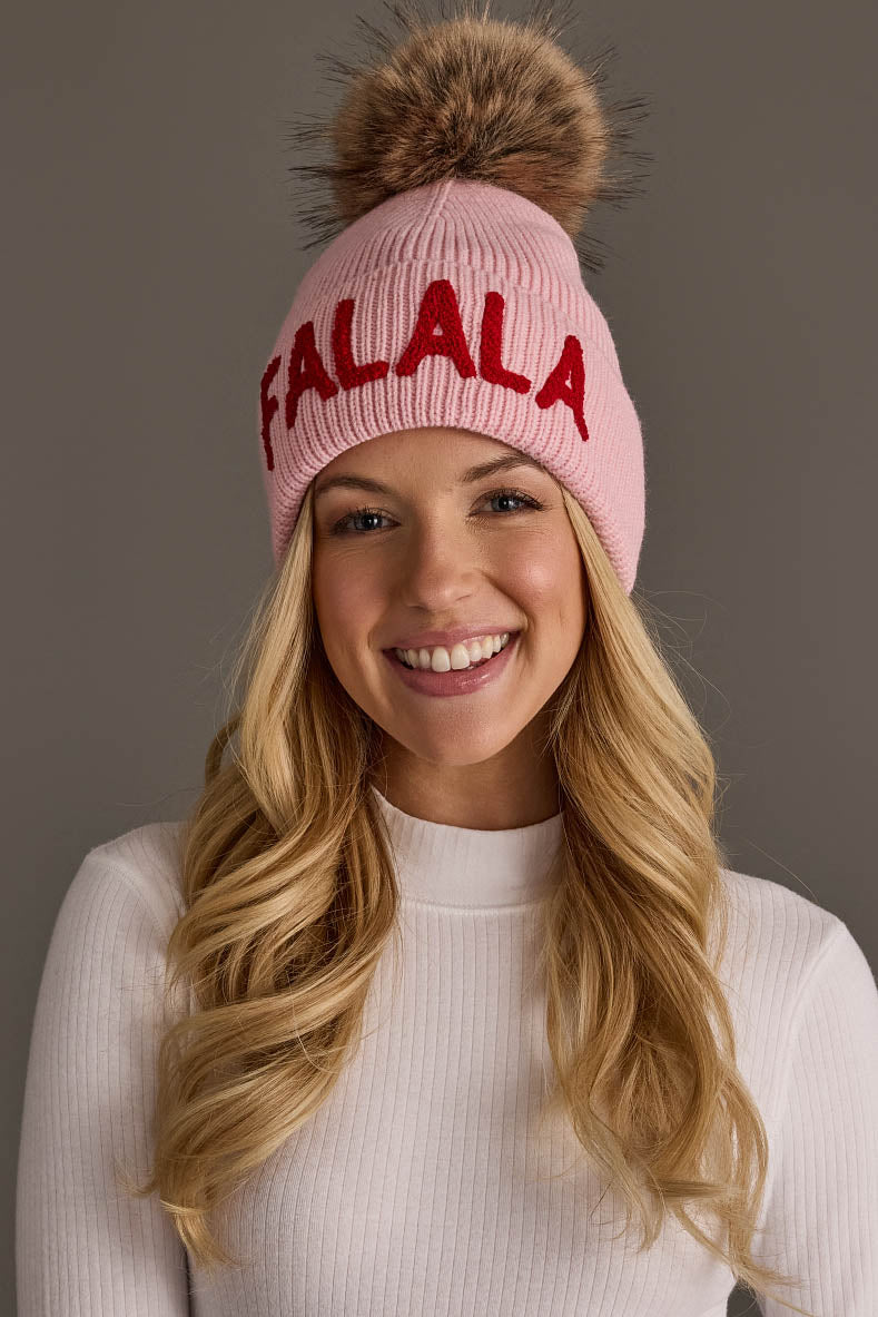 A woman wearing a pink Fa La La Knit Pom Hat with a faux fur pom-pom and the word FALALA embroidered in red.