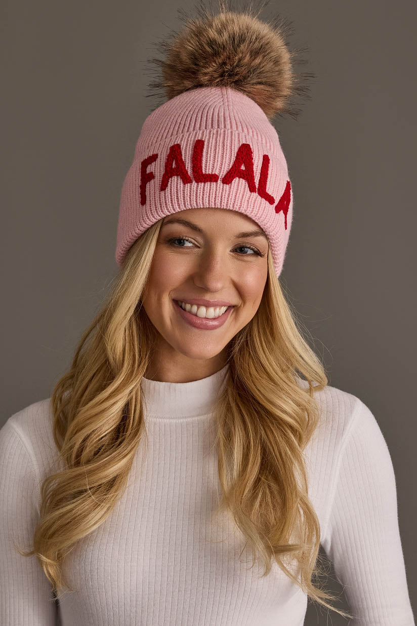 A woman wearing a pink Fa La La Knit Pom Hat with red lettering and a large brown faux-fur pom-pom on top.