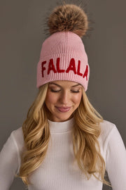 A person wears a Pink Fa La La Knit Pom Hat with red lettering and a brown faux fur pom-pom.