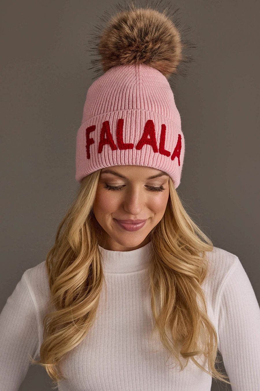A person wears a Pink Fa La La Knit Pom Hat with red lettering and a brown faux fur pom-pom.