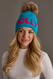 A person wearing a Blue Jolly Knit Pom Hat with pink letters and a large, fluffy faux fur pom-pom on top.