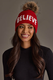 A person wears a Panache Apparel Collection Red Believe Knit Pom Hat, a red knit hat with the word BELIEVE in white letters and a brown faux-fur pom-pom on top.
