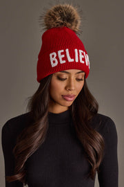 A person wearing a Red Believe Knit Pom Hat, featuring a red ribbed knit design, a white BELIEVE inscription, a fleece lining, and a large faux fur pom-pom on top.