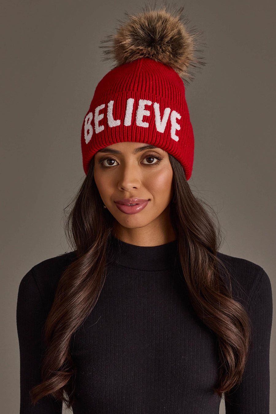 A Red Believe Knit Pom Hat, this fleece-lined red knit beanie features the word BELIEVE in white letters and a large brown faux fur pom-pom on top.