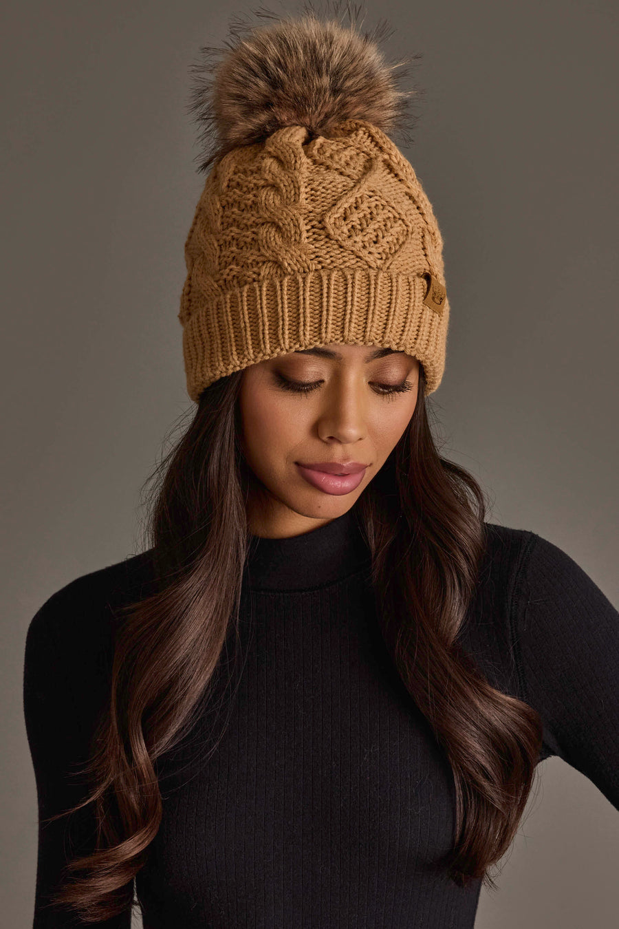 A woman with long dark hair is wearing the Panache Apparel Collection Khaki Cable Knit Pom Hat and a black ribbed turtleneck top, looking down against a plain gray background.