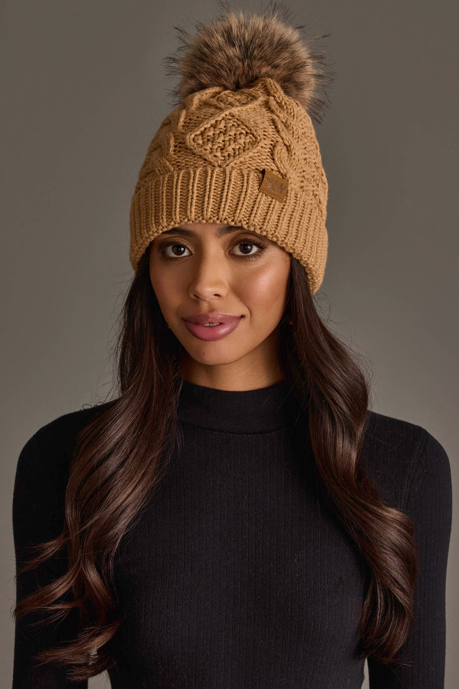 A woman with long, wavy hair stands against a plain gray background, wearing the Panache Apparel Collection’s Khaki Cable Knit Pom Hat and a black ribbed long-sleeve top.