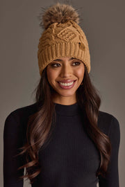 A woman with long brown hair, smiling against a neutral background, wears the Panache Apparel Collection Khaki Cable Knit Pom Hat paired with a black ribbed sweater.