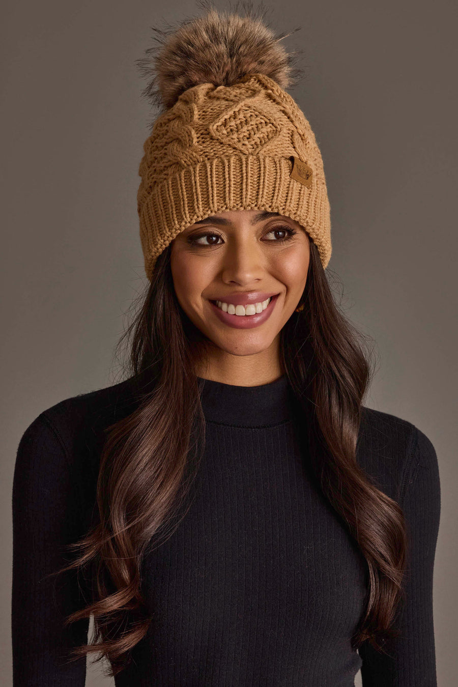 A woman with long brown hair, smiling against a neutral background, wears the Panache Apparel Collection Khaki Cable Knit Pom Hat paired with a black ribbed sweater.