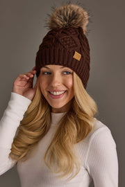 A woman with long blonde hair wears the Dark Brown Cable Knit Pom Hat from Panache Apparel Collection, featuring a faux fur pom-pom. She is smiling and dressed in a white ribbed sweater, posing against a plain background.