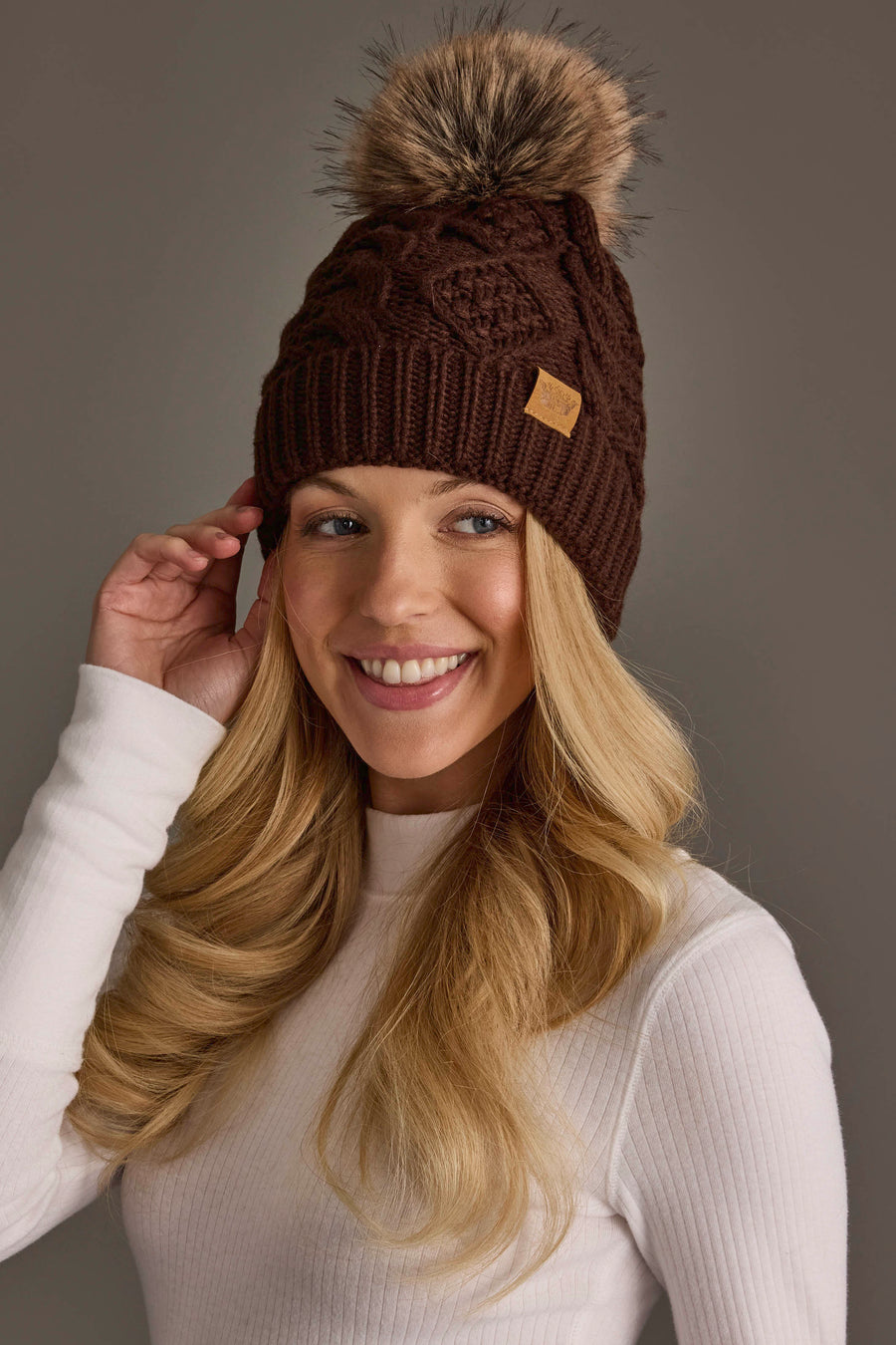 A woman with long blonde hair wears the Dark Brown Cable Knit Pom Hat from Panache Apparel Collection, featuring a faux fur pom-pom. She is smiling and dressed in a white ribbed sweater, posing against a plain background.