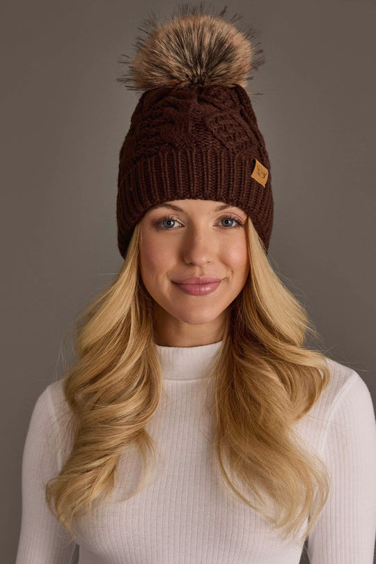 A woman with long blonde hair smiles while wearing the Panache Apparel Collection Dark Brown Cable Knit Pom Hat, featuring a fur pom-pom. She is dressed in a white ribbed top and posed in front of a neutral brown background.