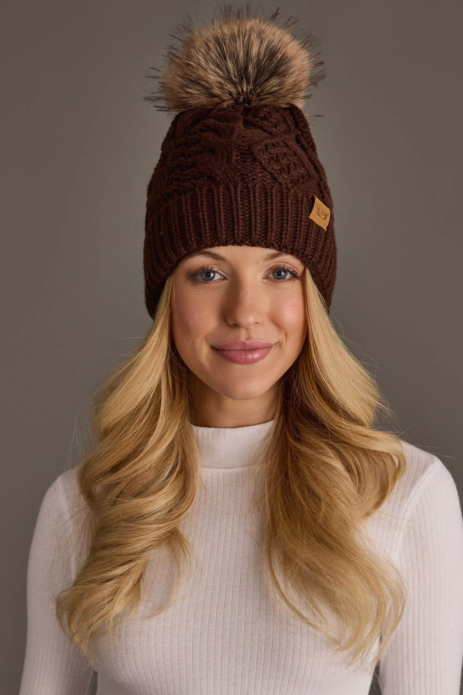 A woman with long blonde hair smiles while wearing the Panache Apparel Collection Dark Brown Cable Knit Pom Hat, featuring a fur pom-pom. She is dressed in a white ribbed top and posed in front of a neutral brown background.