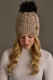 A woman with long blonde hair wears the Panache Apparel Collection Warm Taupe Cable Knit Pom Hat and a white ribbed turtleneck, looking down against a plain grey background.