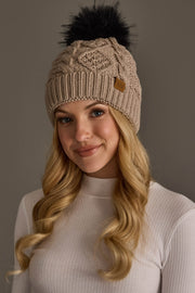 A woman with long blonde hair smiles softly against a neutral background, wearing the Panache Apparel Collection’s Warm Taupe Cable Knit Pom Hat and a light ribbed turtleneck.