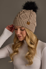 A woman with long blonde hair wearing the Warm Taupe Cable Knit Pom Hat by Panache Apparel Collection and a white ribbed sweater, looking down and adjusting her hat against a grey background.