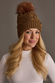 A smiling woman with long blonde hair wears the Panache Apparel Collection Brown Cable Knit Hat with Dark Orange Pom and a white ribbed shirt, standing against a plain gray background.