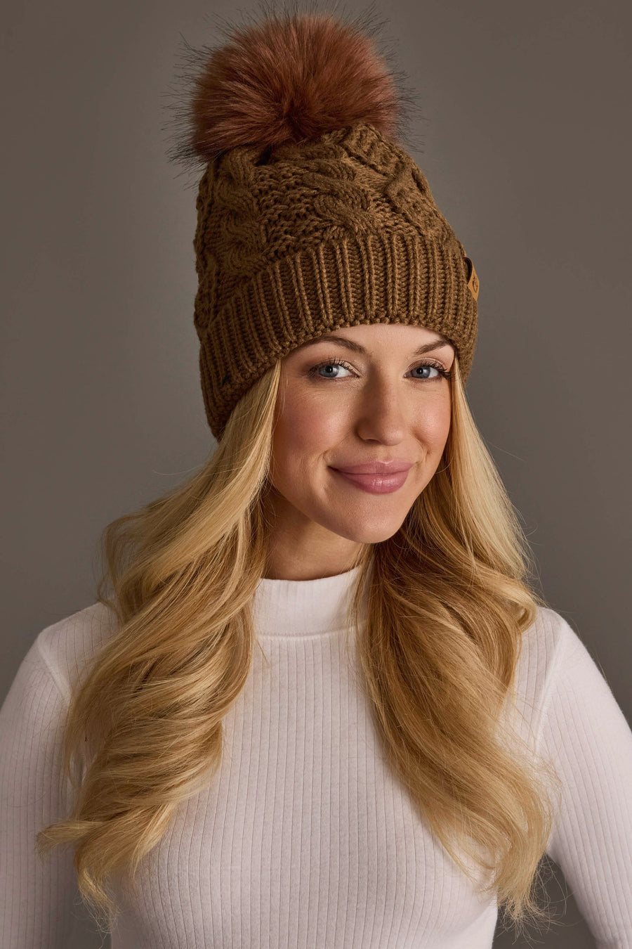 A smiling woman with long blonde hair wears the Panache Apparel Collection Brown Cable Knit Hat with Dark Orange Pom and a white ribbed shirt, standing against a plain gray background.