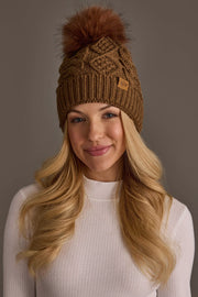 A woman with long blonde hair smiles softly against a plain brown background, wearing the Panache Apparel Collection Brown Cable Knit Hat with Dark Orange Pom and a white ribbed top.