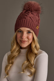 A person with long blonde hair is wearing the Panache Apparel Collection Dark Pink Cable Knit Hat with Dark Pink Pom and a light ribbed turtleneck sweater, smiling softly against a plain gray background.