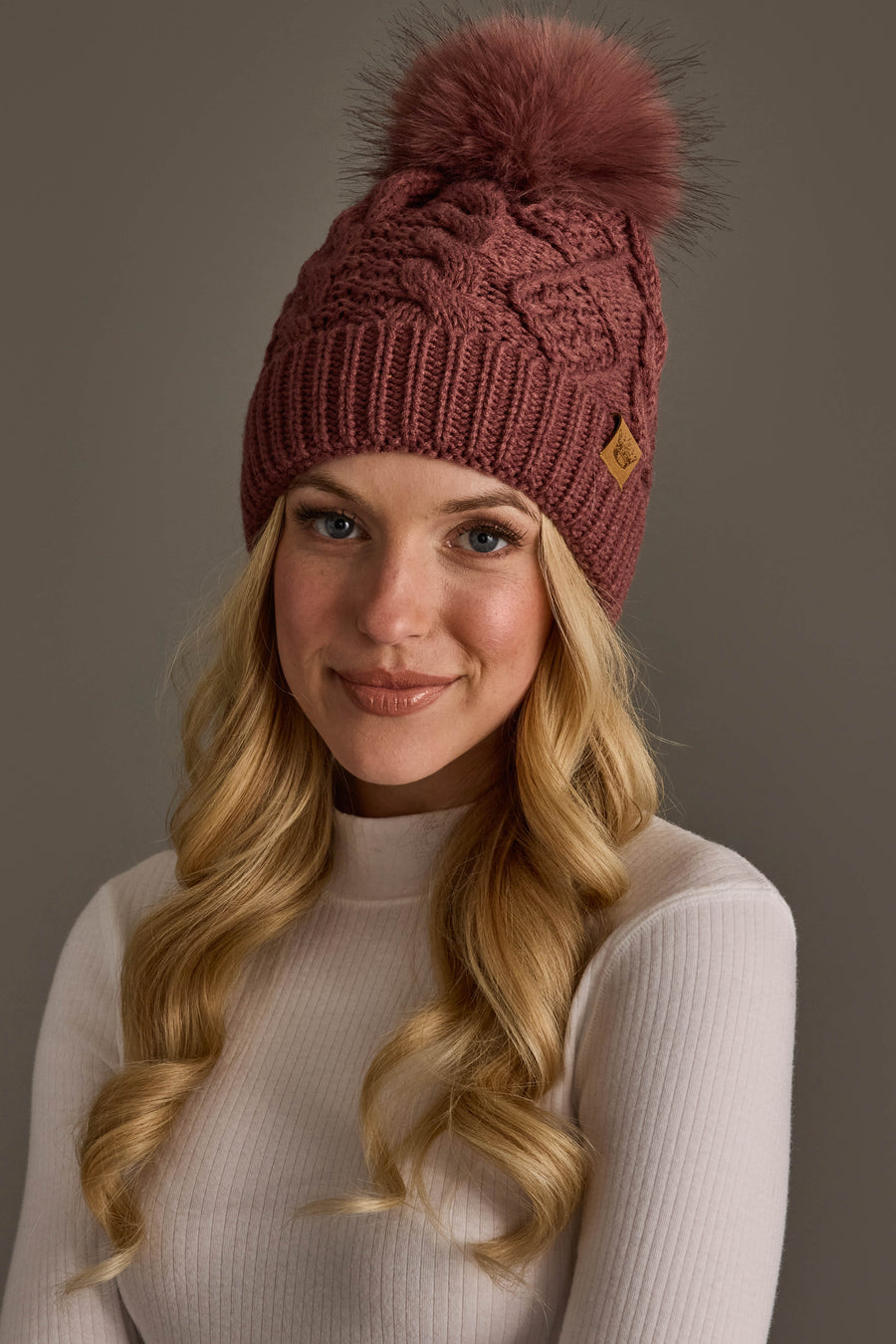 A person with long blonde hair is wearing the Panache Apparel Collection Dark Pink Cable Knit Hat with Dark Pink Pom and a light ribbed turtleneck sweater, smiling softly against a plain gray background.