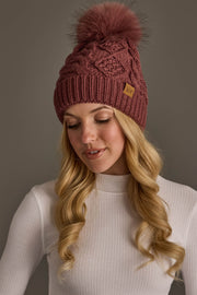 A woman with long blonde hair wears the Panache Apparel Collection Dark Pink Cable Knit Hat with Dark Pink Pom and a fitted white ribbed turtleneck top, looking down and smiling slightly.