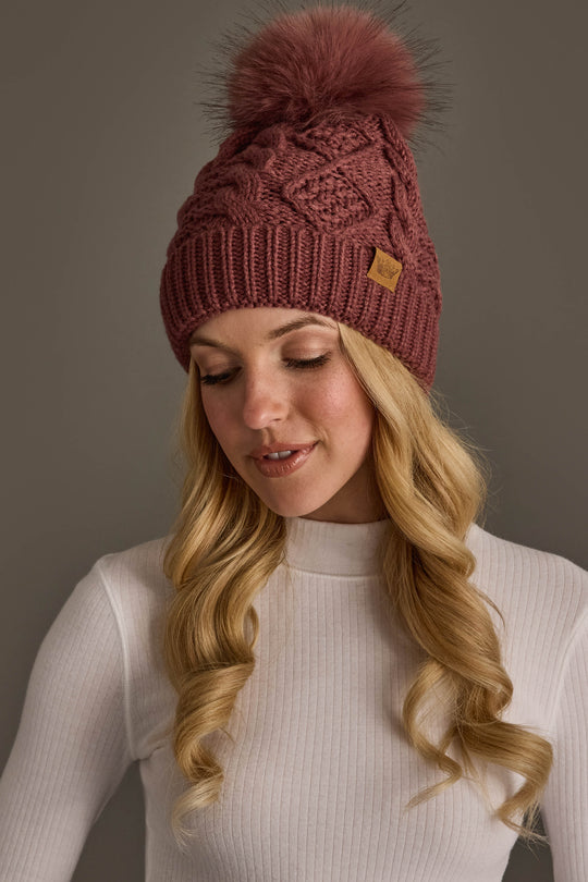 A woman with long blonde hair wears the Panache Apparel Collection Dark Pink Cable Knit Hat with Dark Pink Pom and a fitted white ribbed turtleneck top, looking down and smiling slightly.