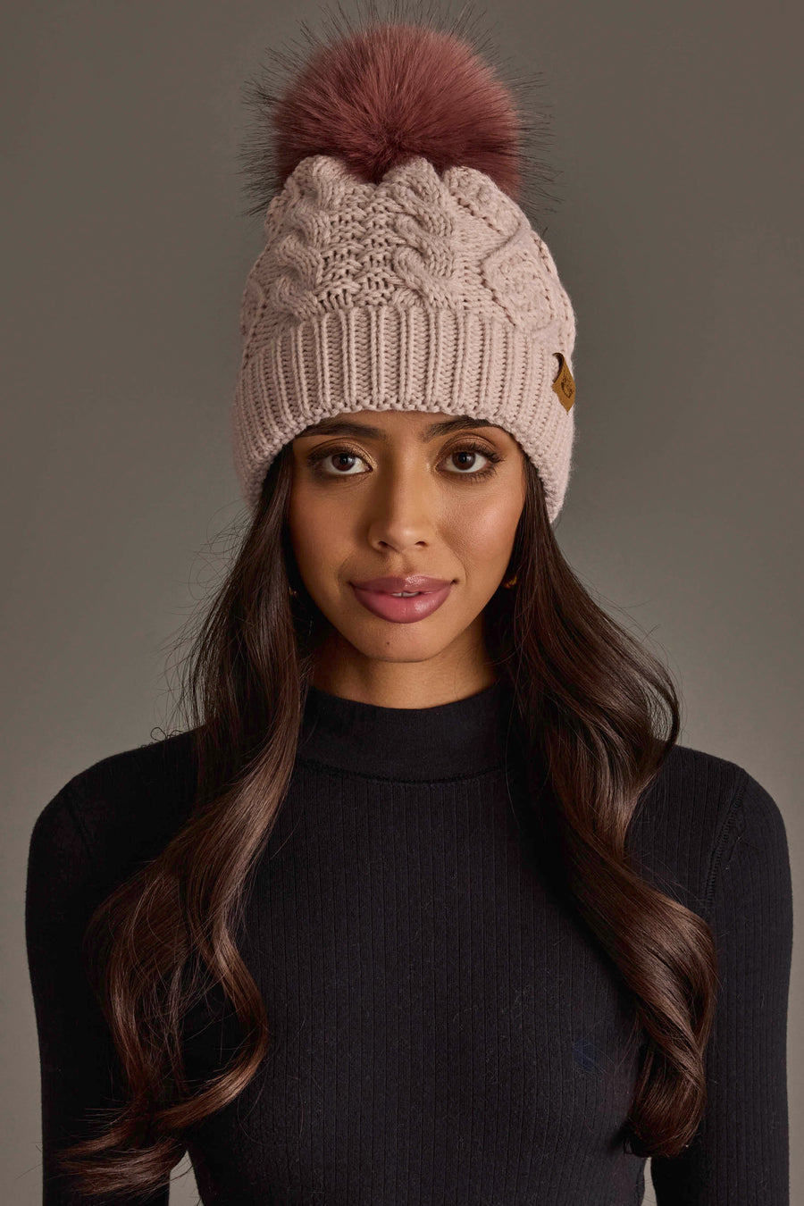 A woman with long brown hair models the Panache Apparel Collection Light Pink Cable Knit Hat with Dark Pink Pom and a black ribbed sweater, posing against a plain gray background.