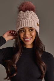 A smiling woman with long brown hair wears the Panache Apparel Collection’s Light Pink Cable Knit Hat with Dark Pink Pom and a black ribbed sweater, posing against a plain gray background.