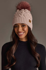 A woman smiles while wearing the Panache Apparel Collection’s Light Pink Cable Knit Hat with Dark Pink Pom, paired with a black long-sleeve top and set against a neutral background.