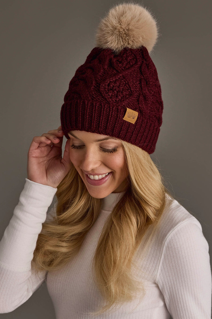 A smiling woman in a white ribbed turtleneck sweater wears the Panache Apparel Collection’s Burgundy Cable Knit Hat with Light Tan Pom, standing against a plain gray background.