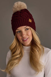 A woman with long blonde hair wears the Panache Apparel Collection’s Burgundy Cable Knit Hat with Light Tan Pom and a white ribbed turtleneck sweater, smiling softly against a plain background.