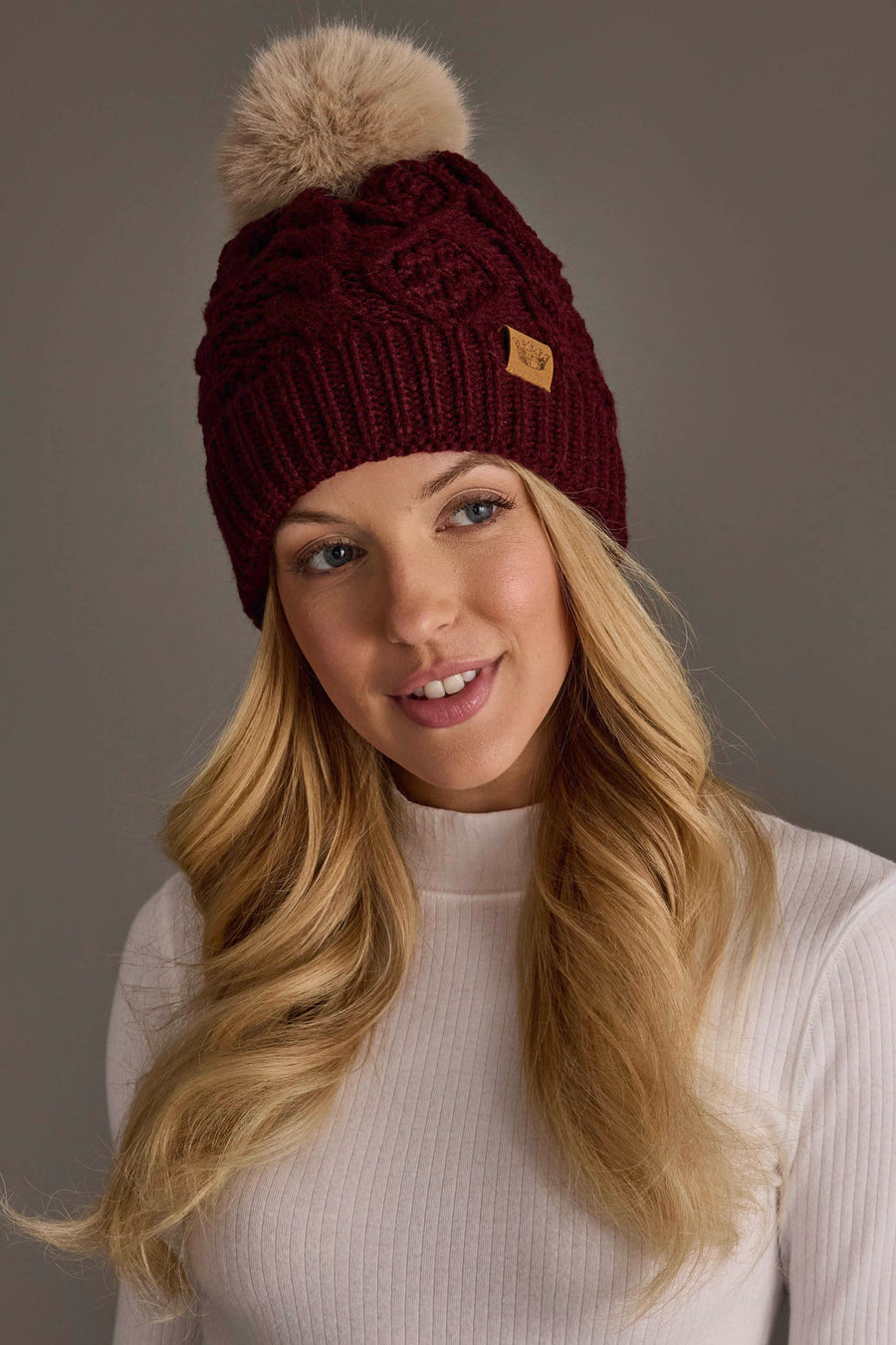 A woman with long blonde hair wears the Panache Apparel Collection’s Burgundy Cable Knit Hat with Light Tan Pom and a white ribbed turtleneck sweater, smiling softly against a plain background.