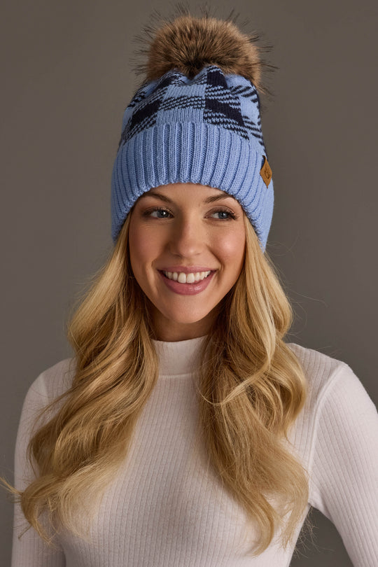 A smiling woman with long blonde hair wears the Panache Apparel Collection Light Blue & Navy Plaid Knit Pom Hat, paired with a white ribbed sweater, standing against a neutral background.
