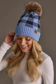 A smiling woman with long blonde hair wears the Panache Apparel Collection Light Blue & Navy Plaid Knit Pom Hat, featuring a large faux fur pom-pom, along with a white ribbed turtleneck sweater, posing against a neutral background.