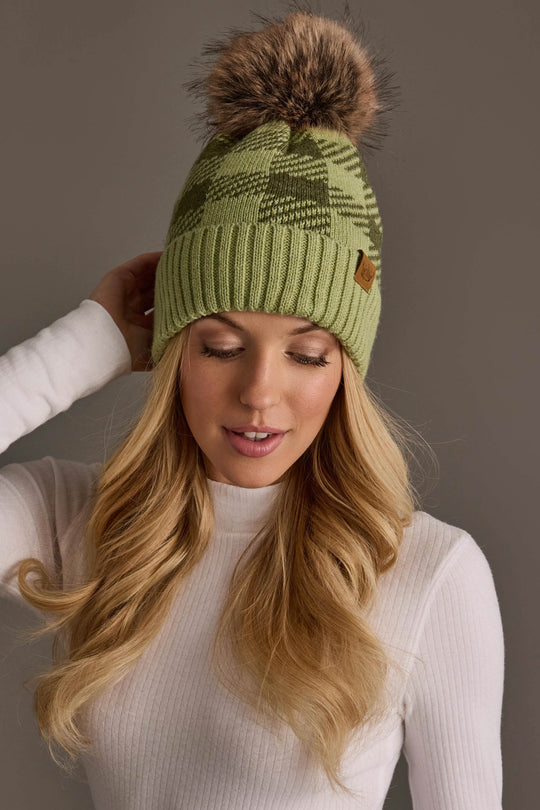 A woman with long blonde hair wears the Panache Apparel Collection Light Green & Olive Plaid Knit Pom Hat and a white ribbed long-sleeve shirt, standing against a neutral background.