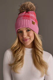 A woman with long blonde hair is wearing the Panache Apparel Collection Pink & Bright Pink Plaid Knit Pom Hat, featuring a brown pom-pom, along with a white ribbed top. She smiles softly against a plain gray background.