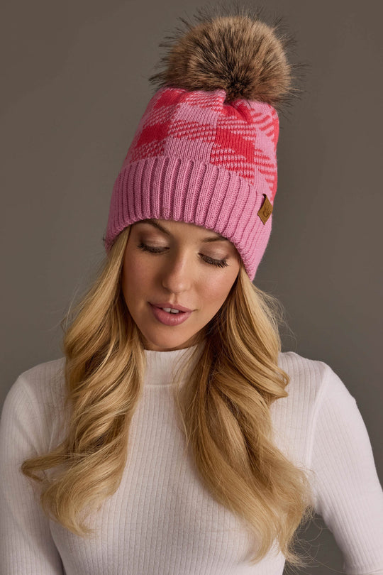 A woman with long blonde hair wears the Pink & Bright Pink Plaid Knit Pom Hat from Panache Apparel Collection and a white ribbed turtleneck sweater, looking down with a neutral expression against a plain gray background.