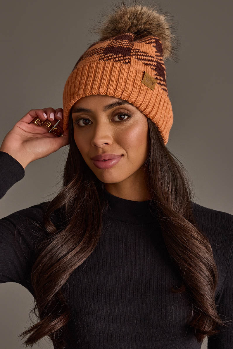A woman wearing the Panache Apparel Collection Orange & Brown Plaid Knit Pom Hat and a black ribbed long-sleeve top poses with one hand touching her hat against a neutral background.