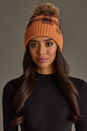 A woman with long brown hair wearing the Panache Apparel Collection’s Orange & Brown Plaid Knit Pom Hat and a black ribbed top poses against a plain gray background.