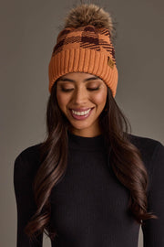 A woman smiles, wearing a black ribbed sweater and the Panache Apparel Collection Orange & Brown Plaid Knit Pom Hat, featuring a faux fur pom-pom, as she stands against a plain gray background.
