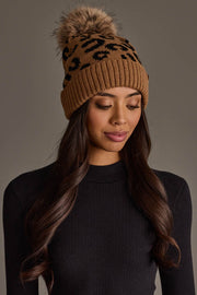 A woman with long wavy hair models the Panache Apparel Collection Camel Leopard Pattern Knit Pom Hat, featuring a brown and black animal print design and a faux fur pom-pom, paired with a ribbed black top, against a plain background.